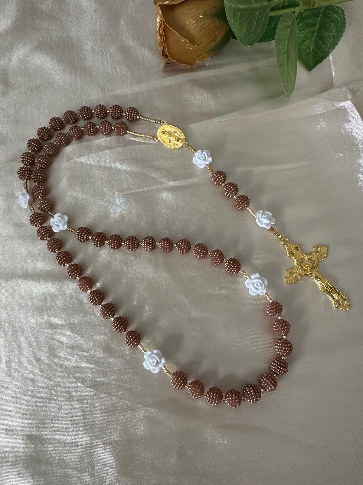 Cocoa Grace Handmade Rosary with textured brown beads, white rose-shaped accents, and gold-tone Virgin Mary centerpiece and crucifix.