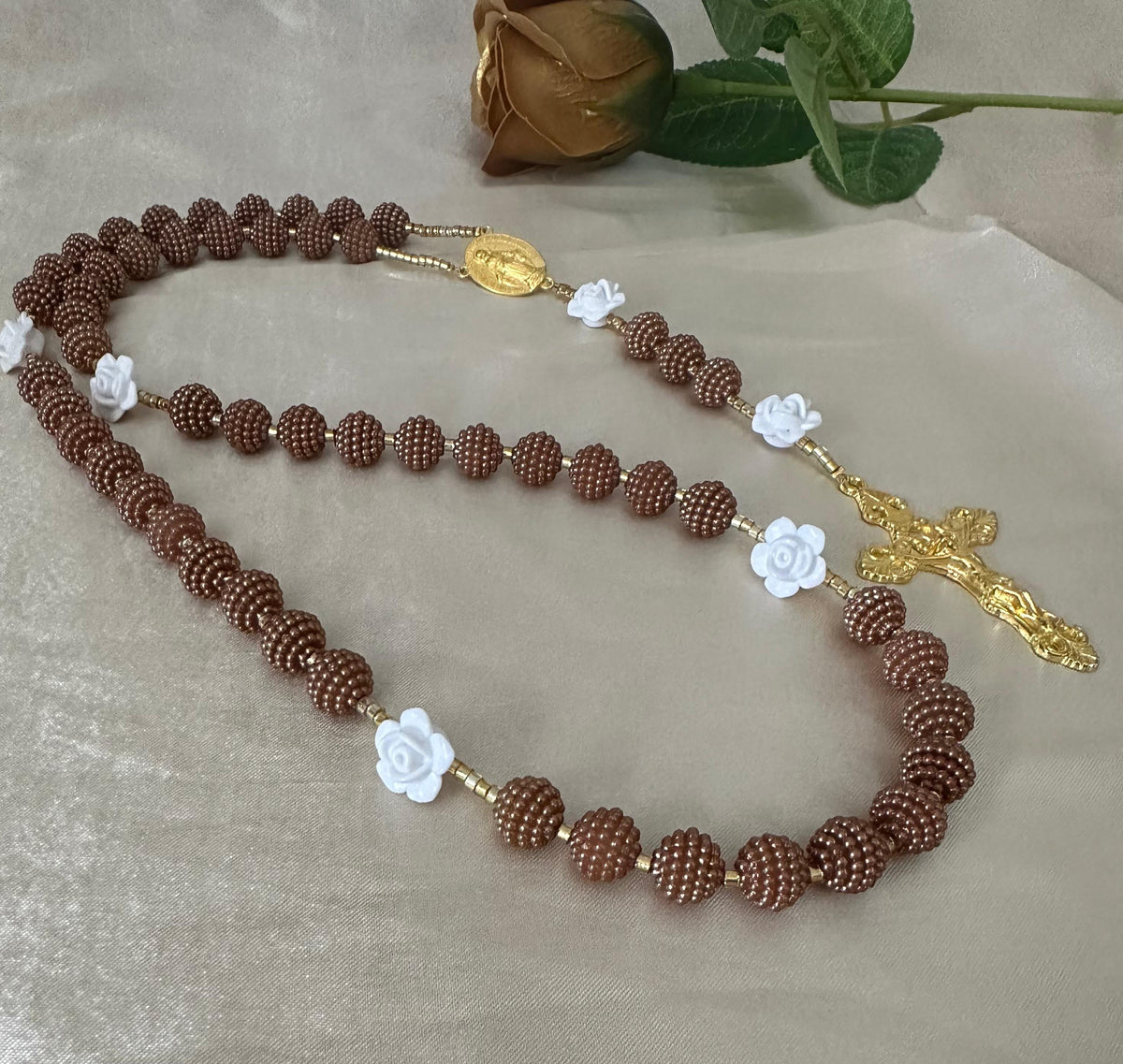 Cocoa Grace Handmade Rosary with textured brown beads, white rose-shaped accents, and gold-tone Virgin Mary centerpiece and crucifix.